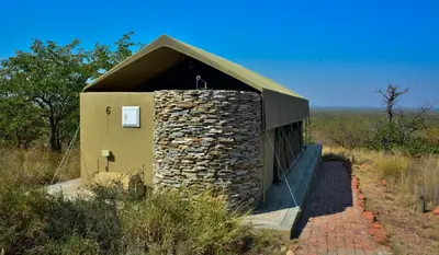 a small building with a stone wall next to a field at Mapesu Wilderness Tented Camp in Musina