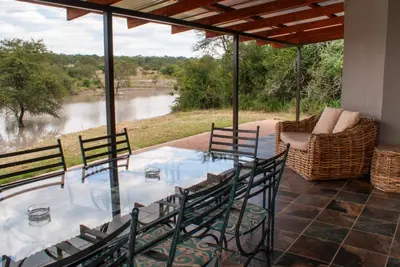 a glass table and chairs on a patio with a view of a river at Casa de la Presa 6 in Polokwane