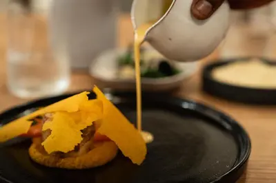 a person pouring sauce on a sandwich on a black plate at Few and Far Luvhondo in Waterpoort
