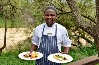a man in an apron holding two plates of food at Rhino Post Safari Lodge in Skukuza