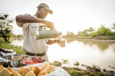 a man preparing food on a table next to a river at Kambaku Safari Lodge in Timbavati Game Reserve