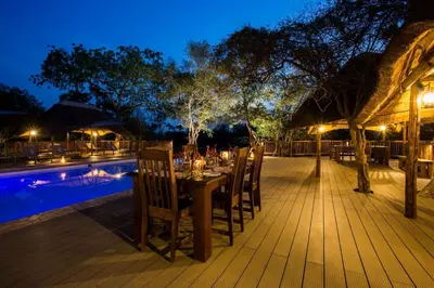 a dining table and chairs next to a pool at night at Vuyani River Lodge in Hoedspruit