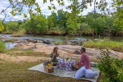 a man and woman sitting on a picnic blanket next to a river at Oase by 7 Star Lodges - Greater Kruger Private 530ha Reserve in Hoedspruit