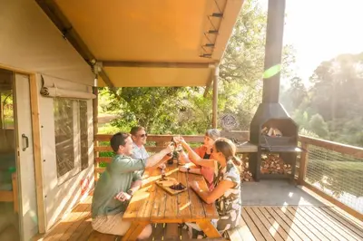 a group of people sitting around a wooden table on a deck with a grill at AfriCamps Magoebaskloof in Magoebaskloof